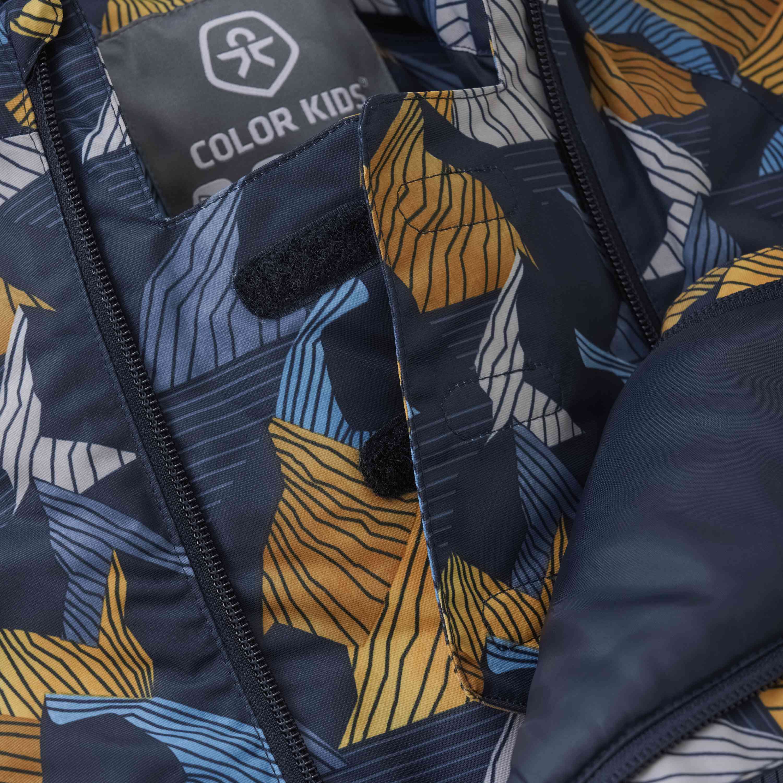 Nahaufnahme einer farbenfrohen Jacke mit abstrakten geometrischen Mustern in Blau-, Gelb- und Weisstönen. Auf dem Stoffetikett im Inneren steht in weisser Schrift „Color Kids“, umgeben von einem stilisierten Kinderlogo. Wir präsentieren den „Color Kids – Overall W. 2 ZIP- AOP“: Die Jacke hat Reissverschlüsse und Klettverschlüsse.
