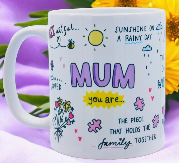 Eine weiße Tasse mit farbenfrohen Illustrationen und herzlichen Botschaften. Auffälliger Text in Pink lautet „MAMA“, umgeben von fröhlichen Zeichnungen von Sonne, Bienen, Blumen und Puzzleteilen. Perfekt als Dekoration fürs Kinderzimmer oder als Ergänzung zu Ihrer Einrichtung fürs Kinderzimmer. Zu den Sätzen gehören „Sonnenschein an einem regnerischen Tag“, „Geliebt“ und „Das Stück, das die Familie zusammenhält“. Dies ist das Feel Good Gift – Tasse Mama von Feel Good Gift.