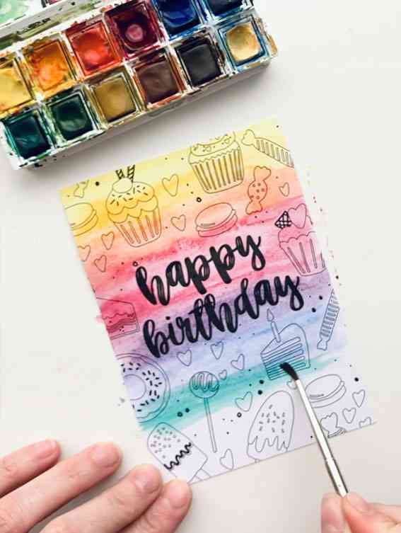 Eine handgemalte Geburtstagskarte zeigt im Hintergrund bunte Aquarellstriche. Die Karte ist mit Kritzeleien von Cupcakes, Herzen und Süßigkeiten verziert. „Happy Birthday“ steht in fetter, schwarzer Schreibschrift. Eine Hand bemalt die Karte mit einem kleinen Pinsel, was an die Schaffung einzigartiger Nynke Ontwerpt – Karte Malkarte Oma von Nynke Ontwerpt erinnert.