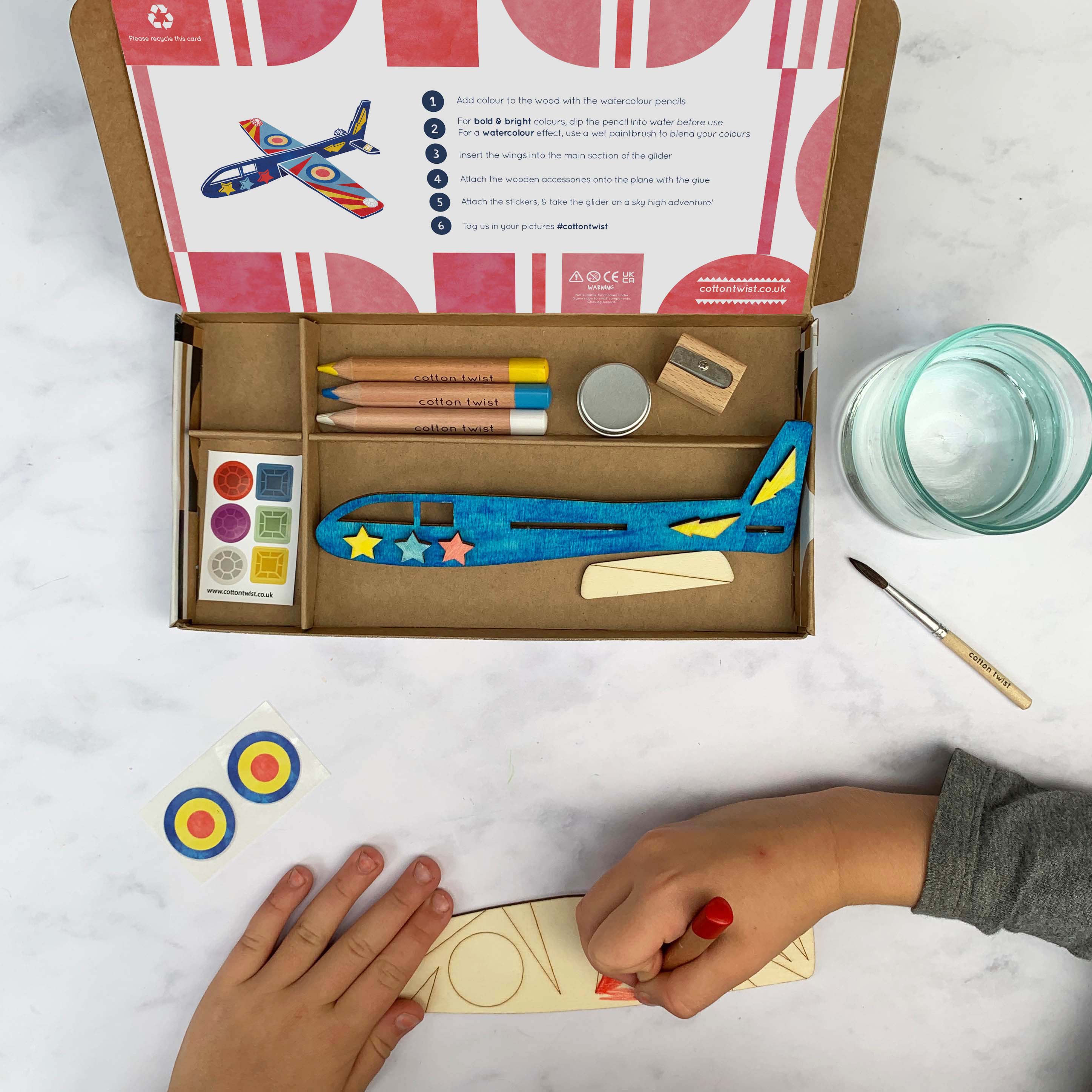 Ein Kind bemalt ein hölzernes Flugzeugmodell mit einem roten Stift aus der **Cotton Twist - DIY Flugzeug-Set Activity Box**. Die Activity Box von **Cotton Twist** enthält Buntstifte, Farbe, Klebstoff und eine ausführliche Anleitung, die auf dem offenen Deckel der Box angezeigt wird. Daneben stehen Wasserkrüge und ein Pinsel. Das fertig bemalte Modell wird ebenfalls als Beispiel gezeigt.