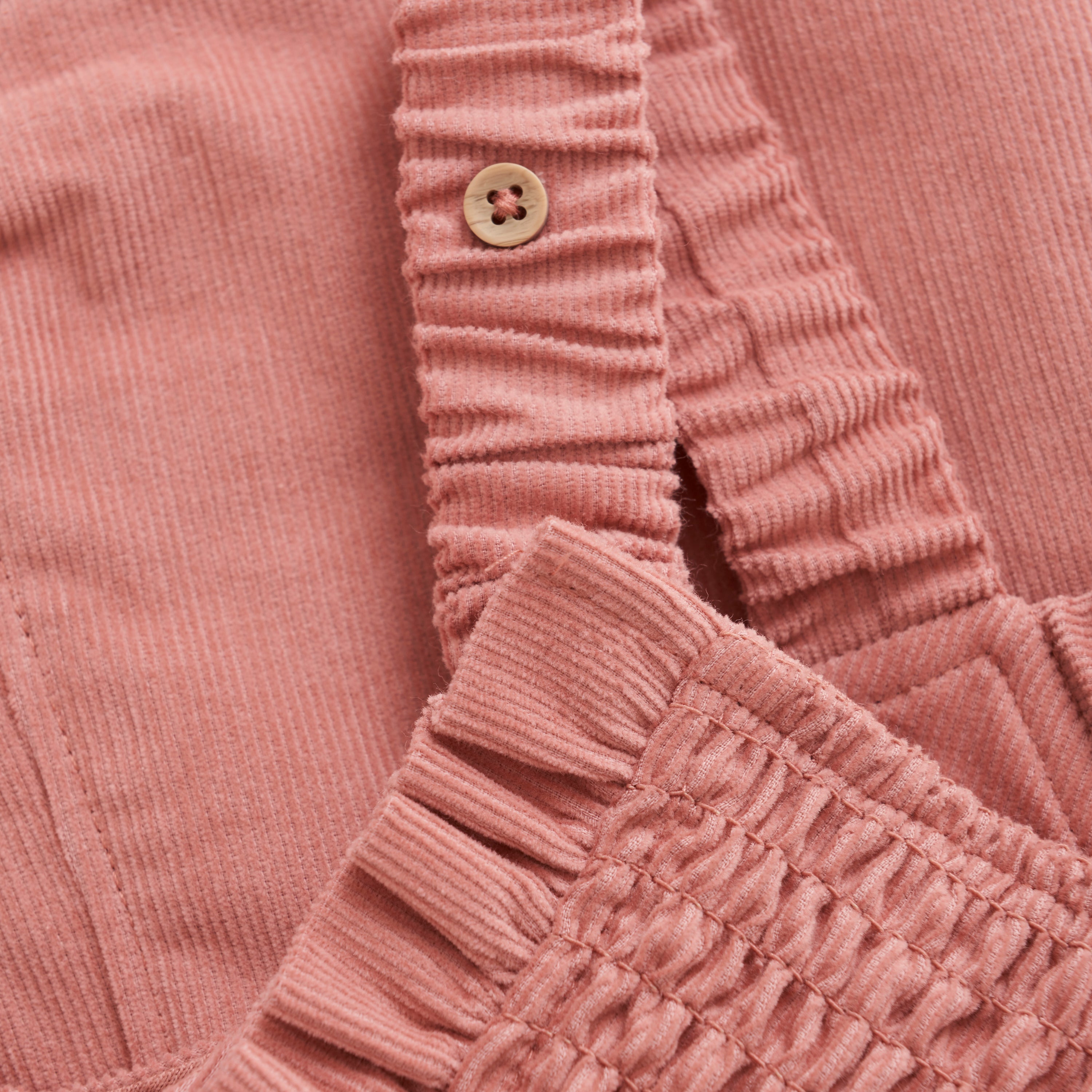 Nahaufnahme des Minymo - Overalls, einem rosa Cord-Kleidungsstück mit einem Bund mit gerafften elastischen Details und einem einzelnen hellbraunen Knopf. Der weiche und gemütliche Stoff besteht aus 100 % Baumwolle und hat eine gerippte Textur. Dieses Stück ist GOTS-zertifiziert, was seine Nachhaltigkeit und Qualität gewährleistet.