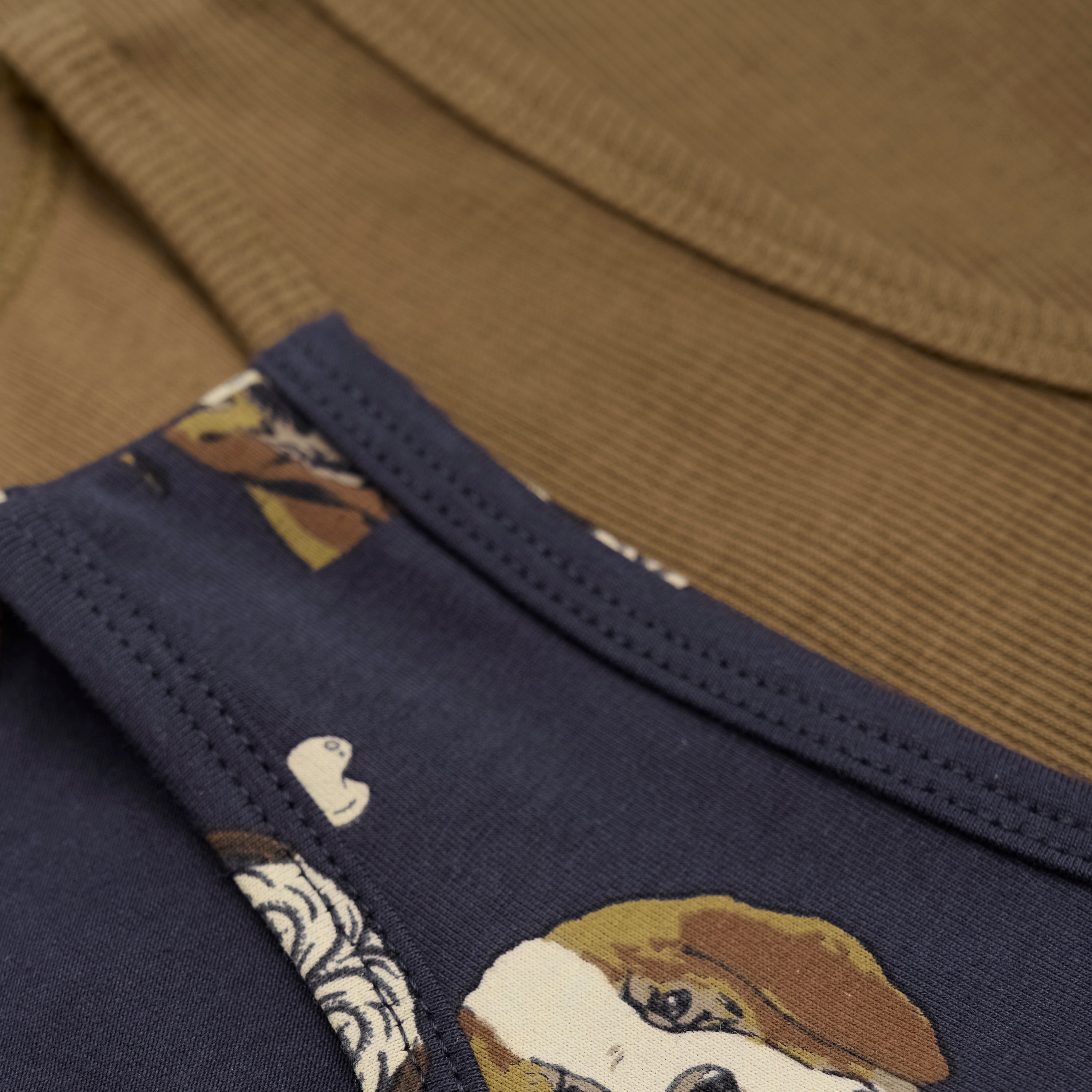Eine Nahaufnahme von zwei gefalteten Minymo - Unterwäsche-Shirts mit sichtbarem Kragen und Ärmelkanten. Das obere Shirt ist marineblau mit einem Aufdruck von braunen und weißen Hunden. Das Shirt darunter ist schlicht braun mit einem strukturierten Stoff, GOTS-zertifiziert für zusätzliche Nachhaltigkeit.
