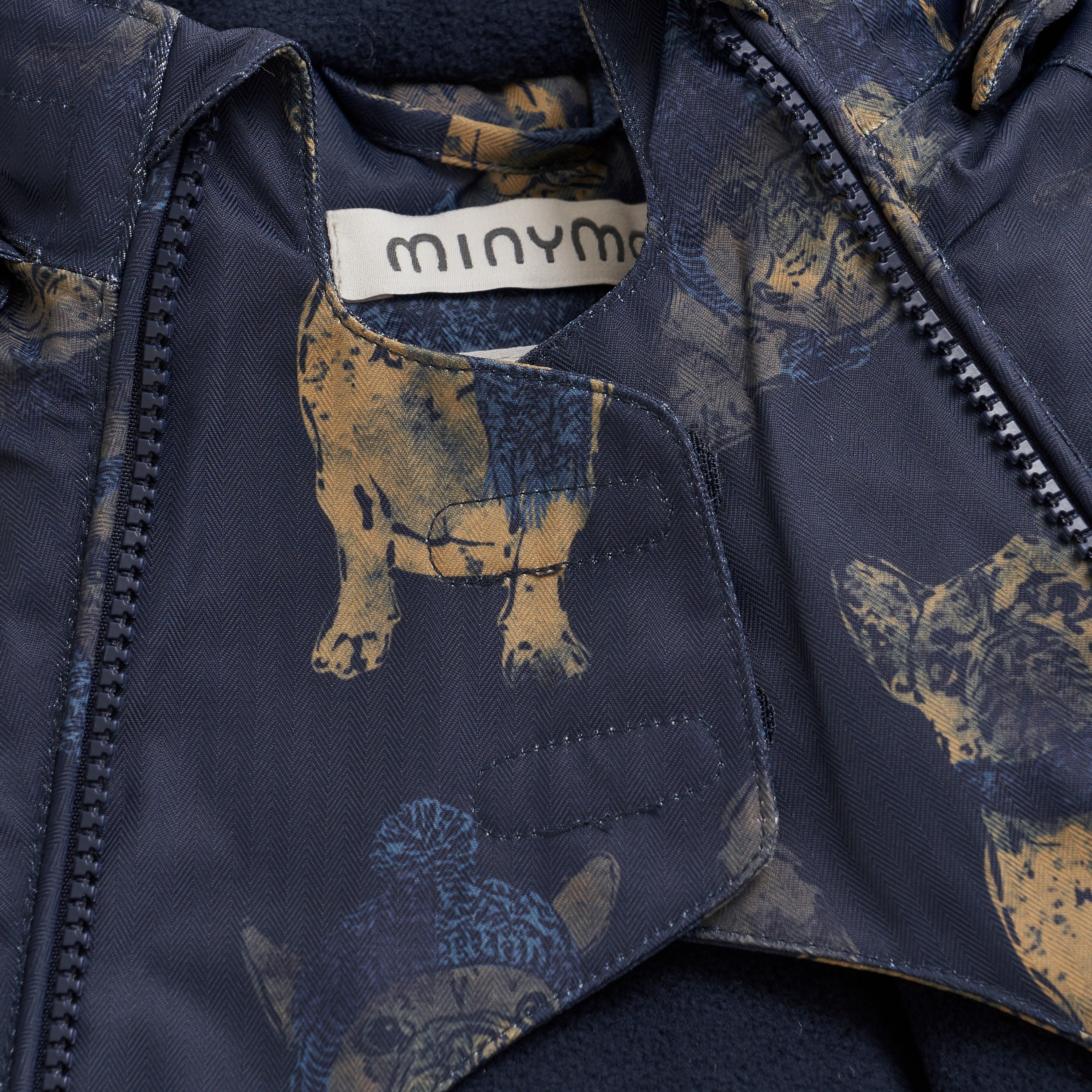 Nahaufnahme der Innenseite eines dunkelblauen Minymo-Schneeanzugs, auf dem ein Etikett mit dem Markennamen „Minymo“ zu sehen ist. Der Stoff ist mit einem Hundemuster in Gelb und Blau bedruckt, von denen einige Hüte tragen. Der Reißverschluss der Jacke ist teilweise geöffnet, sodass der detaillierte Aufdruck auf der Innenseite sichtbar ist. Dieser wärmere Schneeanzug verfügt außerdem über eine abnehmbare Kapuze für vielseitige Verwendung.