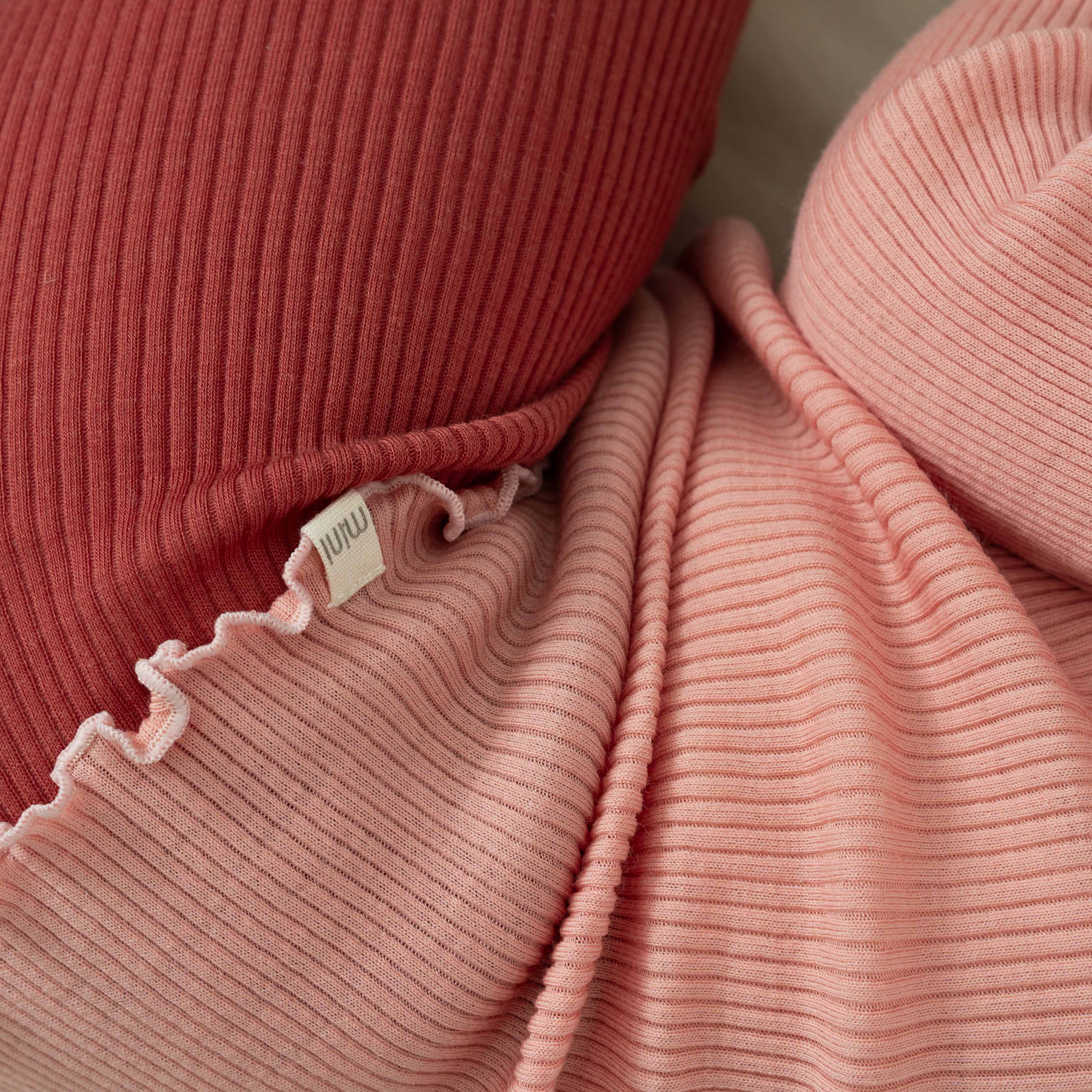 Nahaufnahme von zwei gerippten Strickstoffen in Rosatönen. Der obere Stoff ist ein dunkleres, rötliches Rosa mit einem Rüschenrand, während der untere Stoff ein helleres Rötro ist. Diese Materialien sind weich und strukturiert und eignen sich ideal als Wärmeschicht oder zum Anfertigen des gemütlichen Langarmshirts Minimalisma aus Merinowolle. Ein kleines weißes Etikett ist sichtbar.
