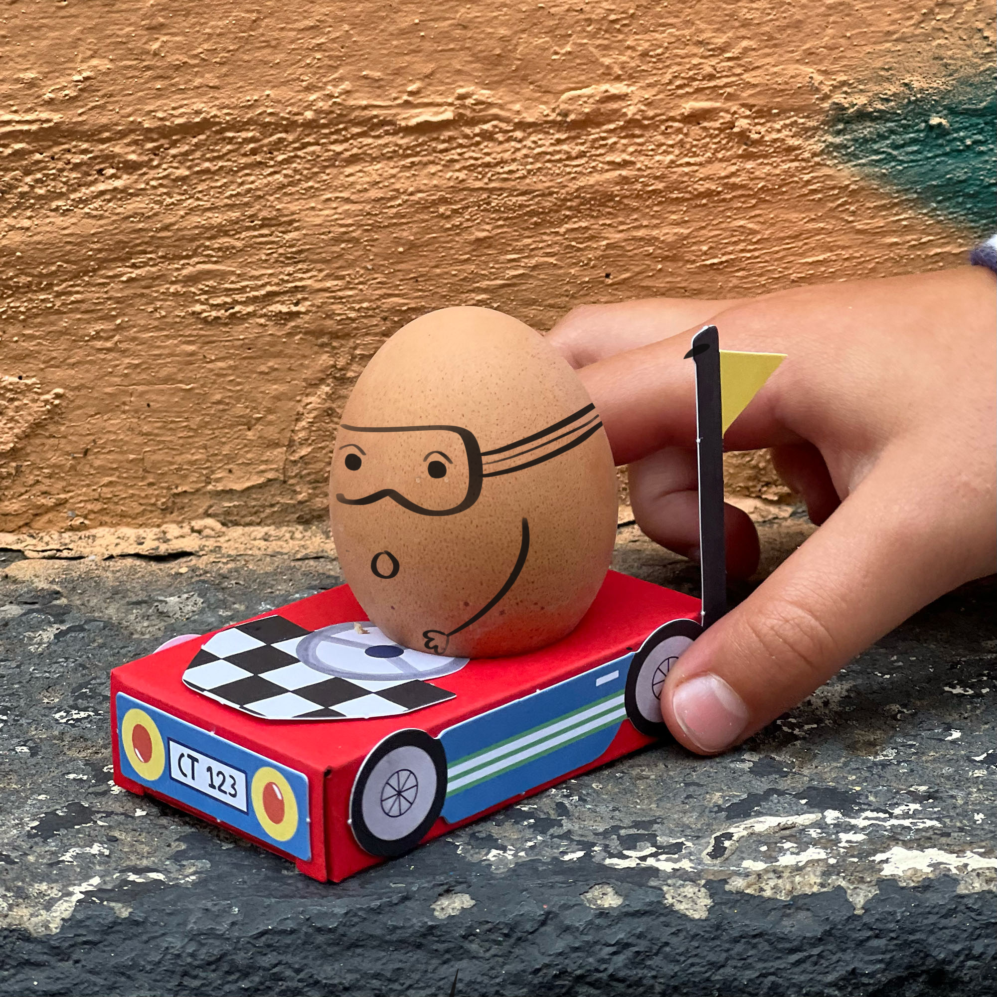 Eine Hand legt ein braunes Ei mit einem gezeichneten Gesicht, einer Schutzbrille und einem Schnurrbart in ein kleines rotes Spielzeugauto aus nachhaltigen Materialien. Das Auto hat ein kariertes Flaggendesign und auf dem Nummernschild steht „CT 123“. Der Hintergrund zeigt eine strukturierte, bemalte Wand. Diese verspielte Szene hebt Cotton Twists DIY Matchbox-Rennwagen der Marke Cotton Twist hervor.