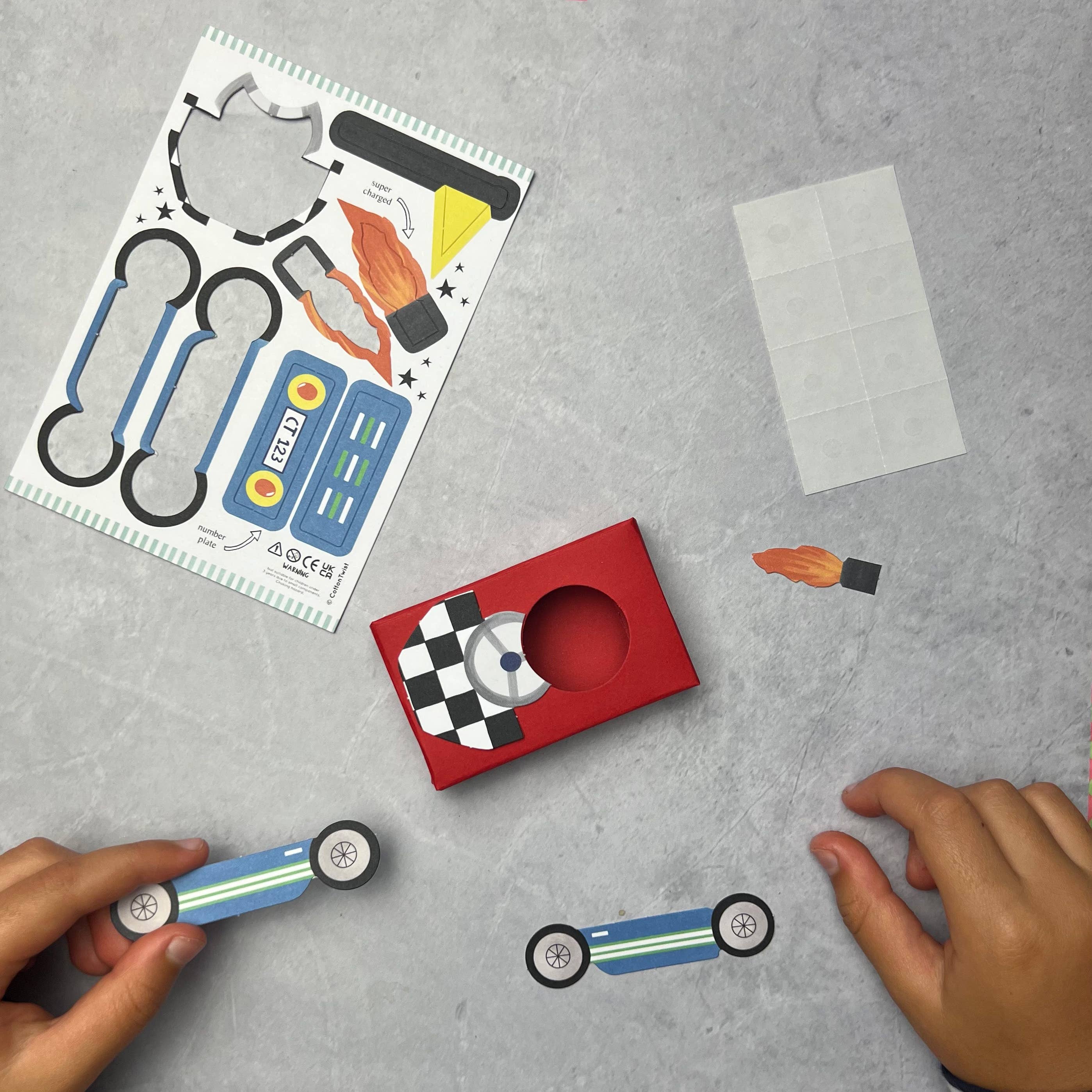 Zwei Hände sind beim Zusammenbau eines Cotton Twist - DIY Matchbox-Rennwagens beteiligt, eine hervorragende Möglichkeit, Feinmotorik zu fördern. Auf dem Tisch liegt ein Blatt mit Papierautoteilen, eine rote Schachtel, eine karierte Flagge und eine kleine Rolle Klebeband. Eine Hand platziert die Karosserie, während die andere das Autogestell mit Rädern hält.